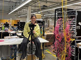 A woman sits behind the camera in a technical room
