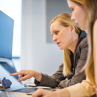 A woman explains something to another on a laptop in an office
