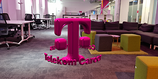 In a office, the T logo floats between furniture as an AR object with a balloon effect in the middle of the room