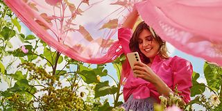 A woman with a pink vail is standing in a flower field. She is holding a smartphone.