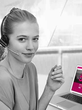 Eine junge Frau mit Headset sitzt im Großraumbüro und lächelt in die Kamera.