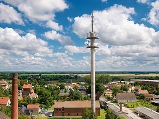 Vor einem blauen Sommerhimmel mit Schäfchenwolken sieht man in einer Luftaufnahme den Mobilfunkmast von Kyritz an der Knatter. 